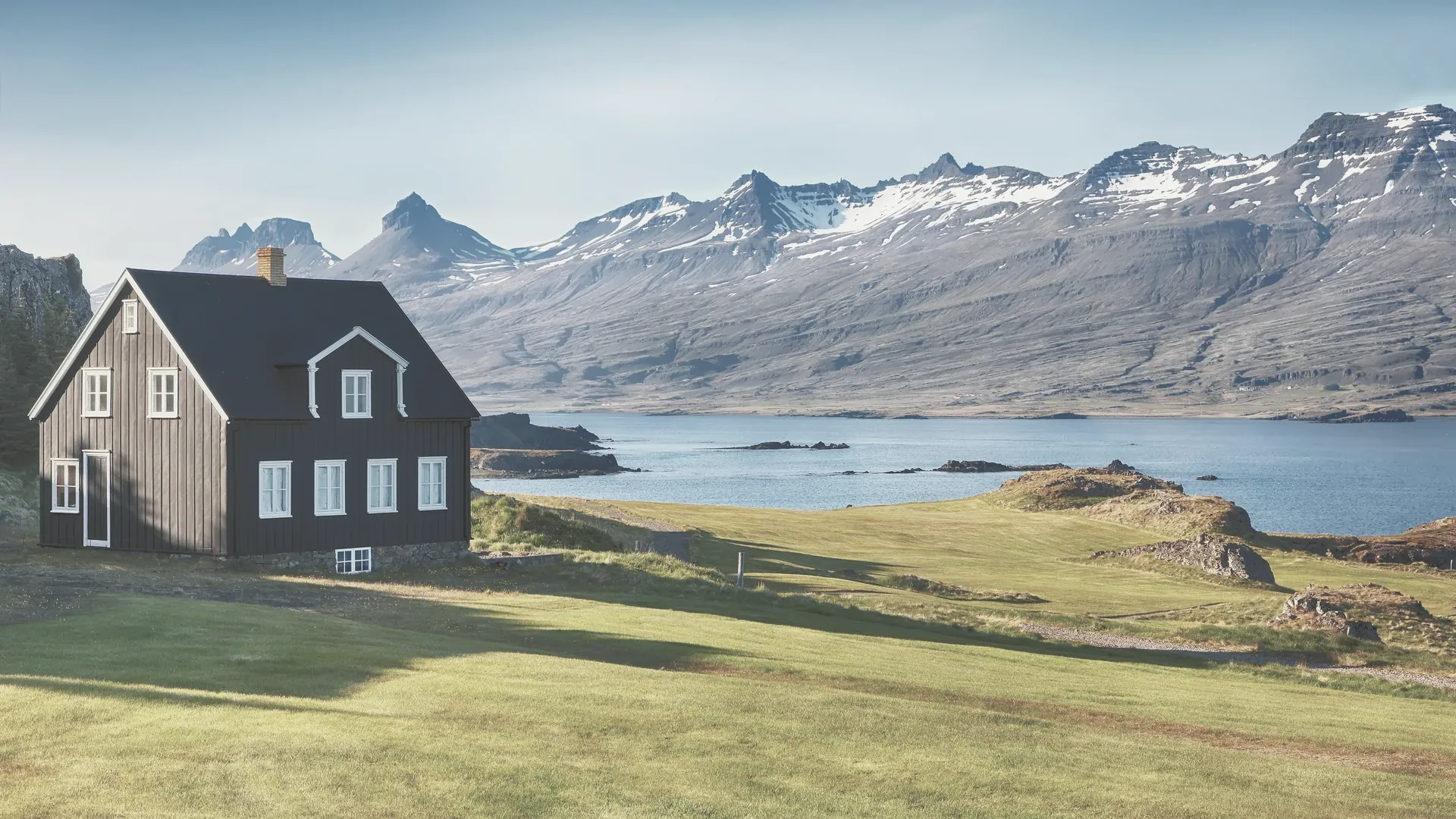 Ein einzelnes Haus am Wasser vor einer Bergkette, im ruhigen Tageslicht